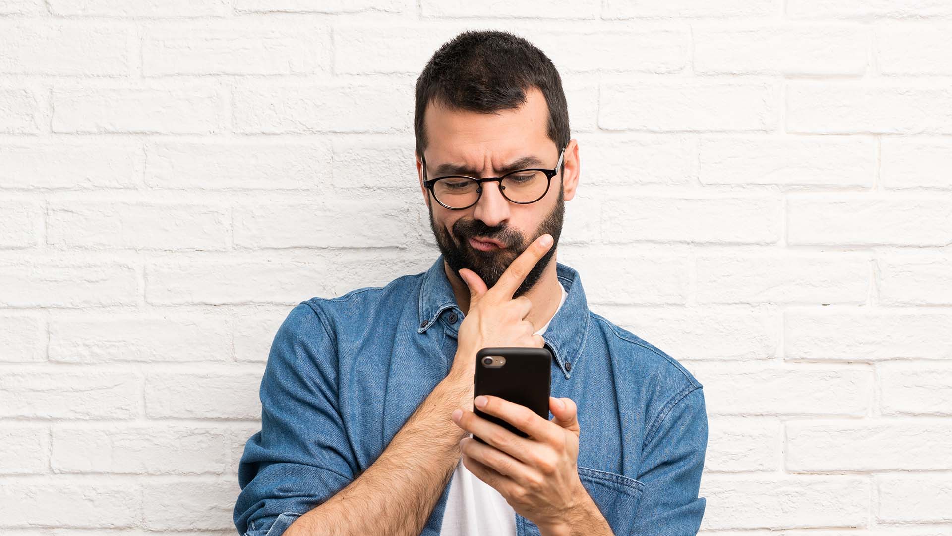Brunette bearded man wearing glasses and jean shirt, rubbing his chin while looking at his phone. White brick background.
